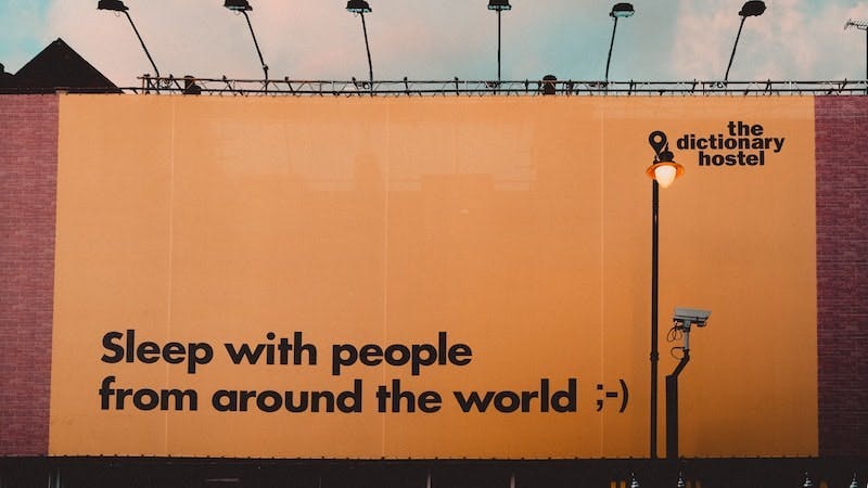 Un panneau publicitaire faisant la promotion de l'auberge Dictionnary, la phrase "Dormir avec des gens du monde entier" est affichée