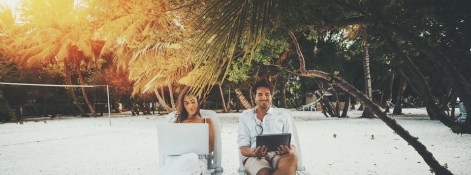 Ihr seht einen Mann und eine Frau auf Liegestühlen im Urlaub am Strand sitzen. Dabei haben Sie ihre Laptops auf dem Schoß.