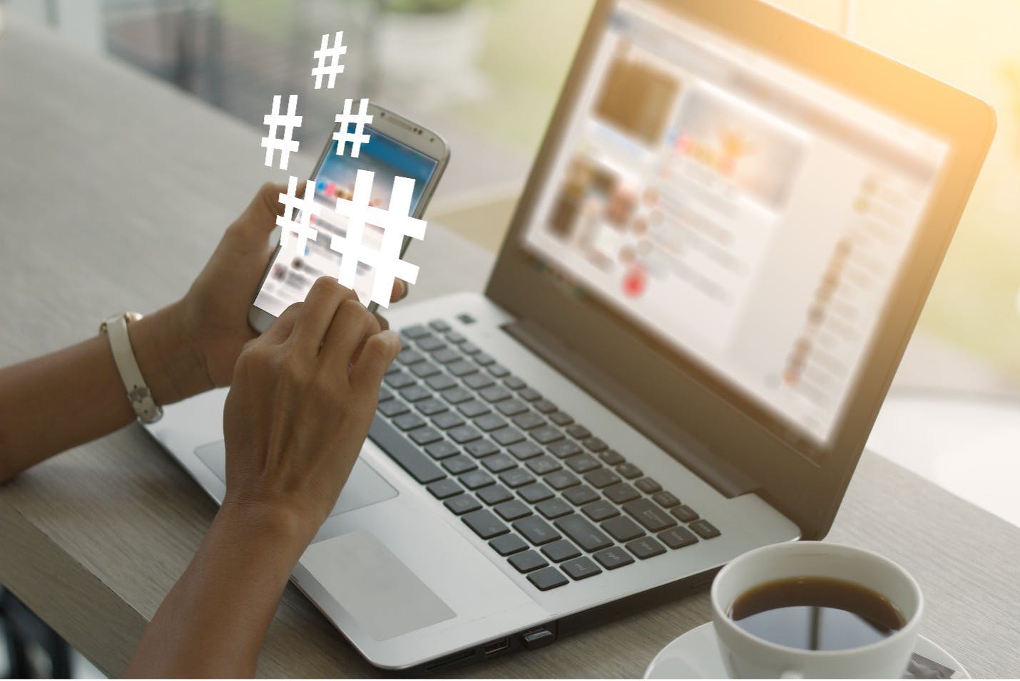 A person sitting in front of a computer looking at a phone that has several hashtag icons hovering above it. Hashtags are a great tool to use to help you find user-generated content for your social wall. 