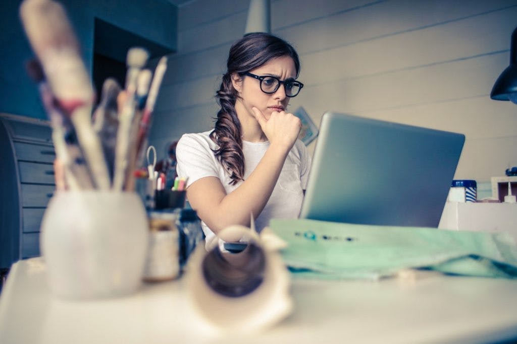 Frau mit Brille sitzt denkend an ihrem Schreibtisch vor ihrem Laptop