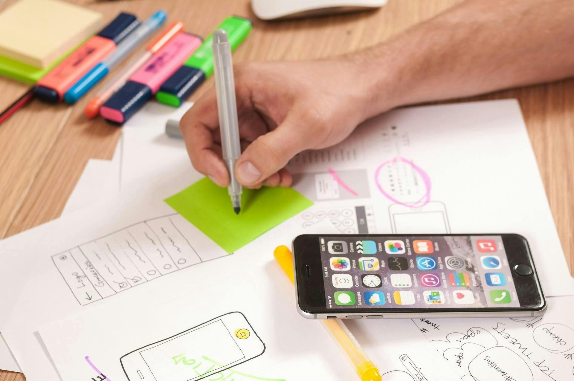 Marketer writing on a green sticky note with an iPhone unlocked showing multiple apps. 