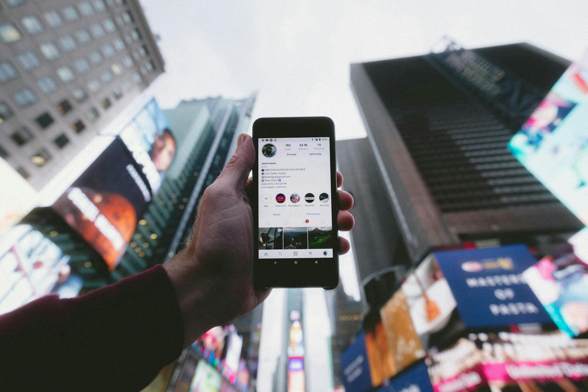 An image of a phone tilted up toward the sky in Times Square of New York City. The phone is opened to the social meida platform Instagram.