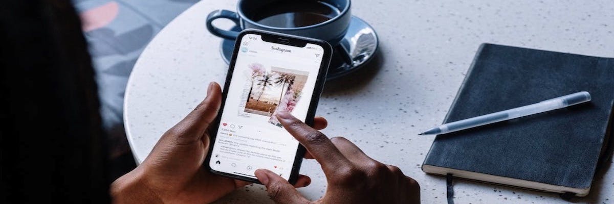 Eine Person hält ein Smartphone in der Hand und stöbert durch Instagram, daneben sind ein Notizbuch und eine Tasse Kaffee zu sehen