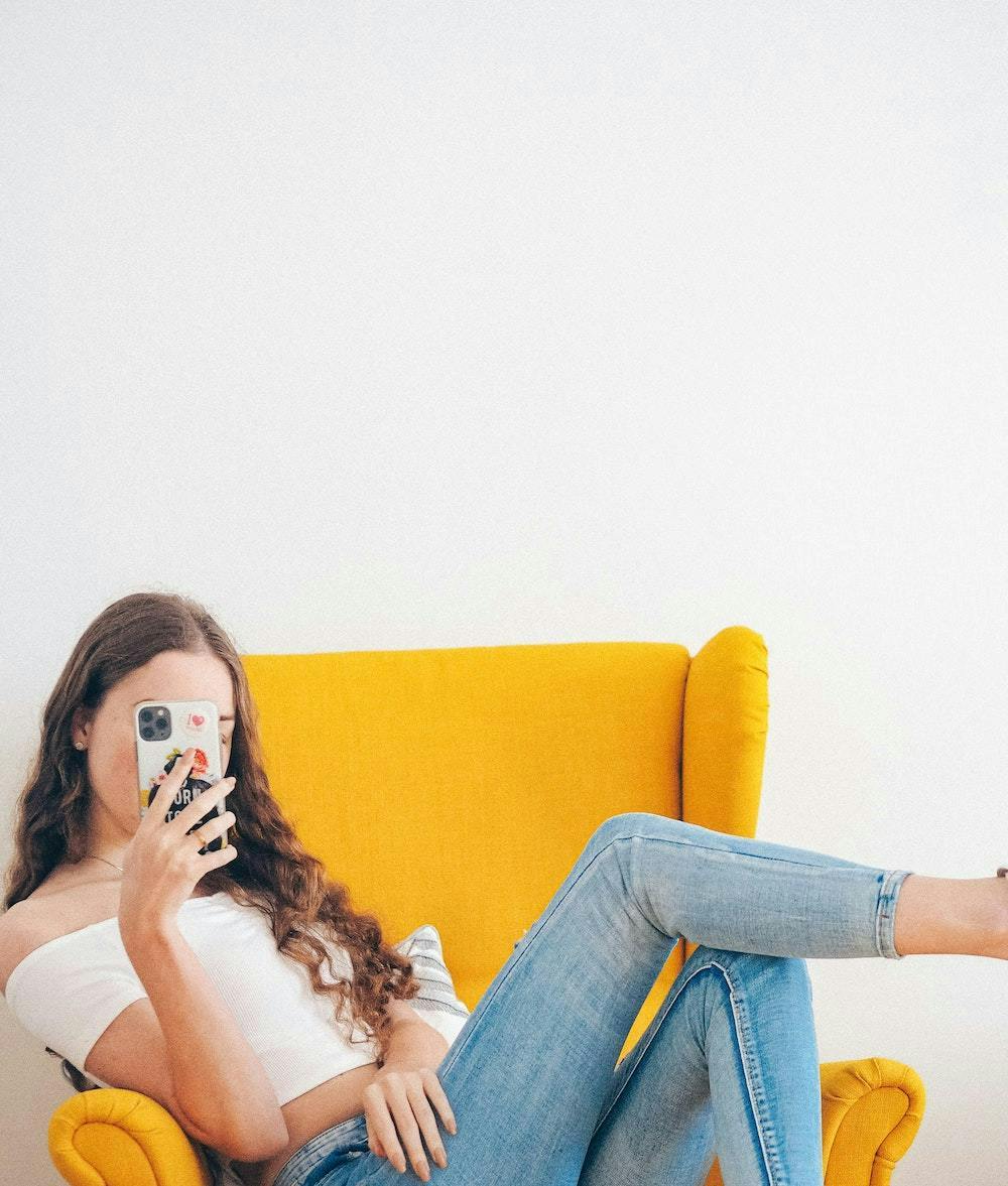 Female influencer sitting in a yellow chair, with face obscured by phone. Fake influencers blog post.