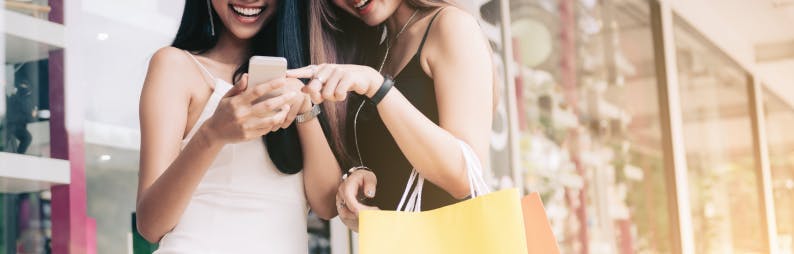 two girls with shopping bags and a cell phone in hand looking at the screen while smiling 