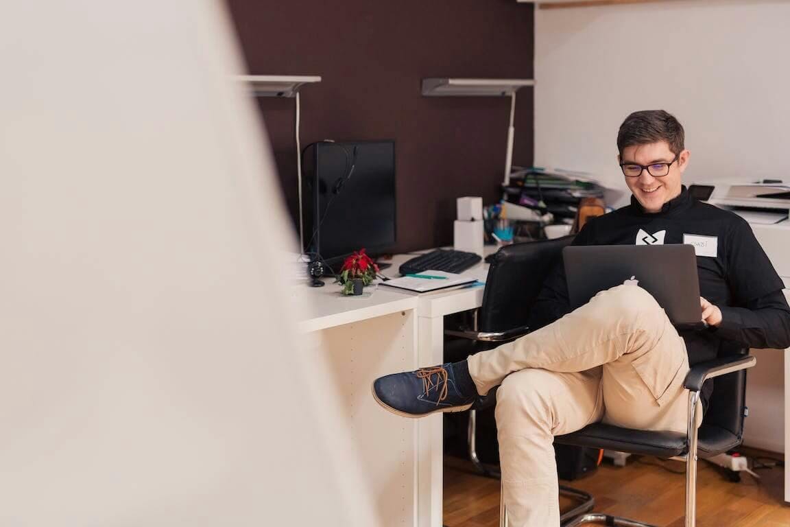 Man sieht einen jungen Mann an seinem Arbeitsplatz sitzen. Er hat die Beine überschlagen und auf seinem Schoß liegt ein Macbook, an dem er arbeitet.