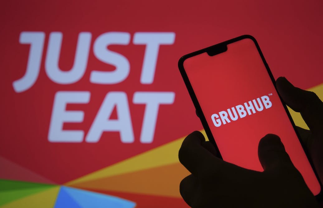 A hand holding a phone with the GrubHub app up, in front of a Just Eat sign 