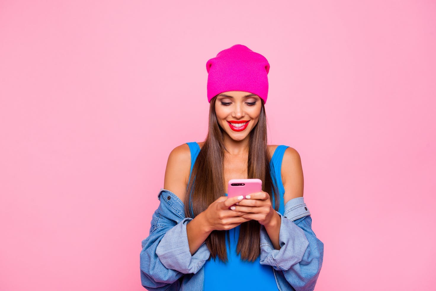 Photo d'une jeune femme qui porte un bonnet rose, un débardeur bleu, une chemise bleue, sourit en regardant son smartphone. Elle se tient contre un mur rose. 