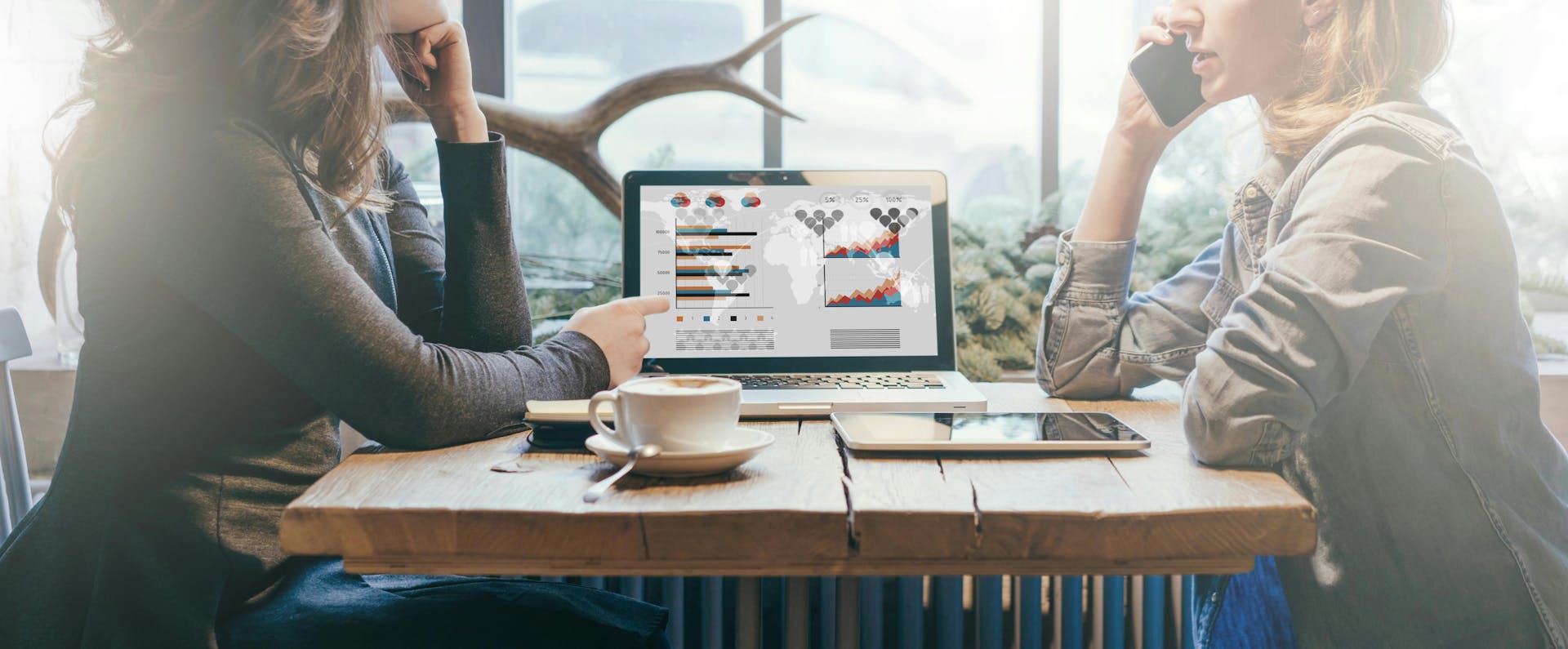 Photo of two women sitting on a desk together while working and drinking coffee