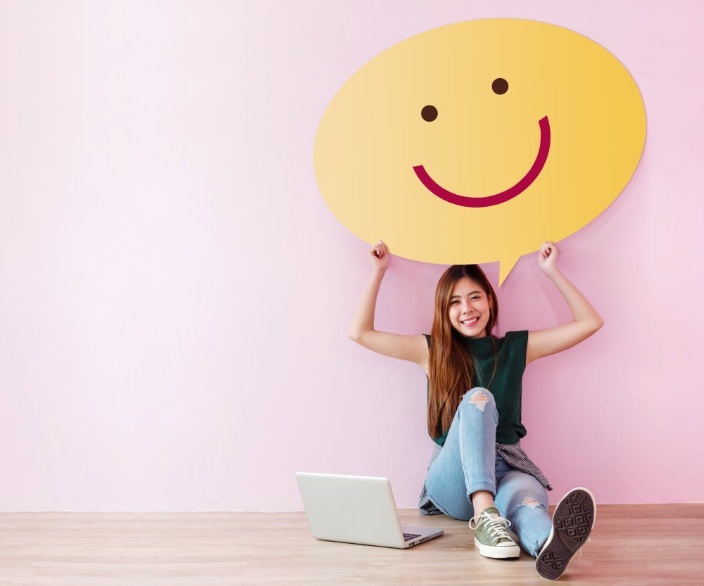 A young gril sitting in front of a laptop holding a large smiley face poster above her head.