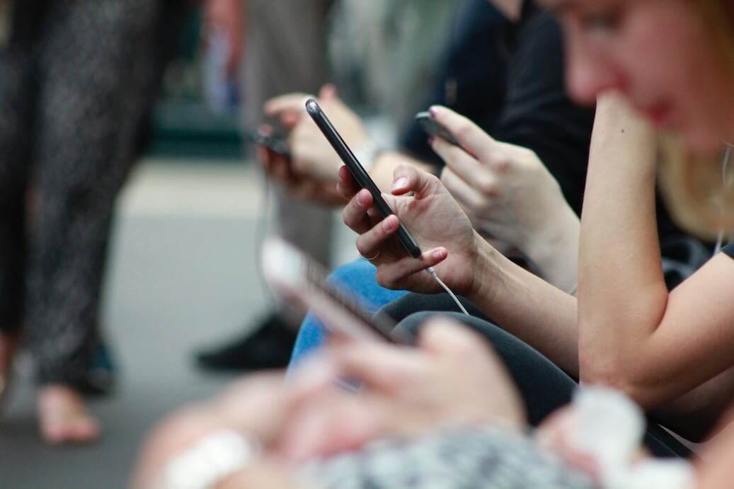 Man sieht ein Foto mehrerer junger Menschen, die in der Stadt sitzen und auf ihre iPhones schauen.
