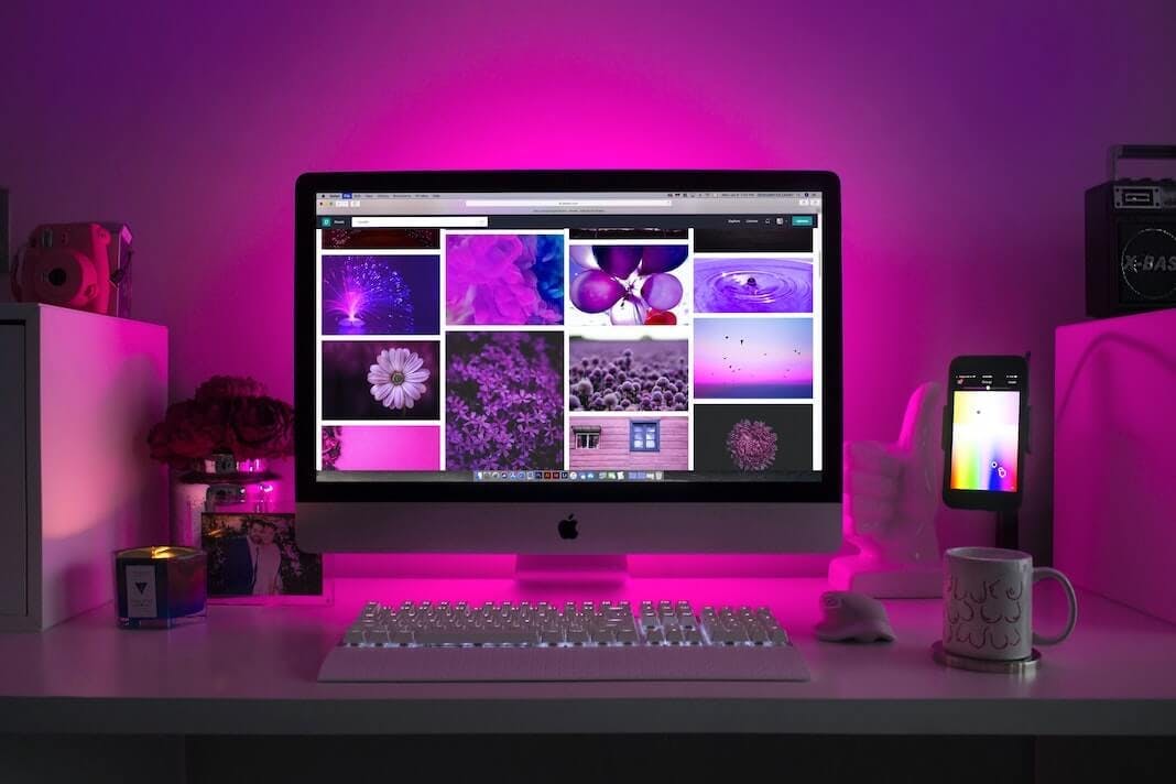Man sieht einen Schreibtisch mit einem Mac, auf dem Kacheln mit verschiedenen Bildern geöffnet sind. Der Max wird von hinten mit lila Licht bestrahlt. Das Bild ist Teil unseres Beitrags zu Social Media Walls.