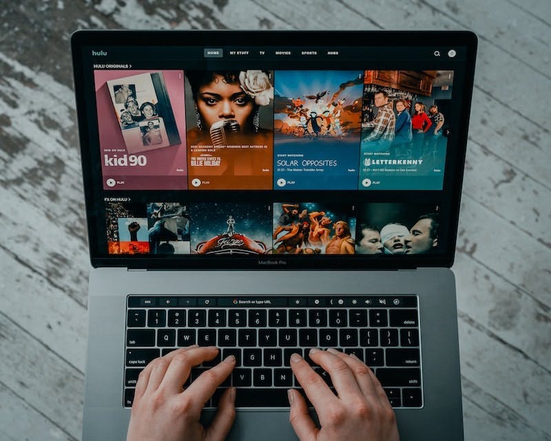 A person is typing on a laptop, on the screen we see the hulu entertainment platform displaying different series and movies