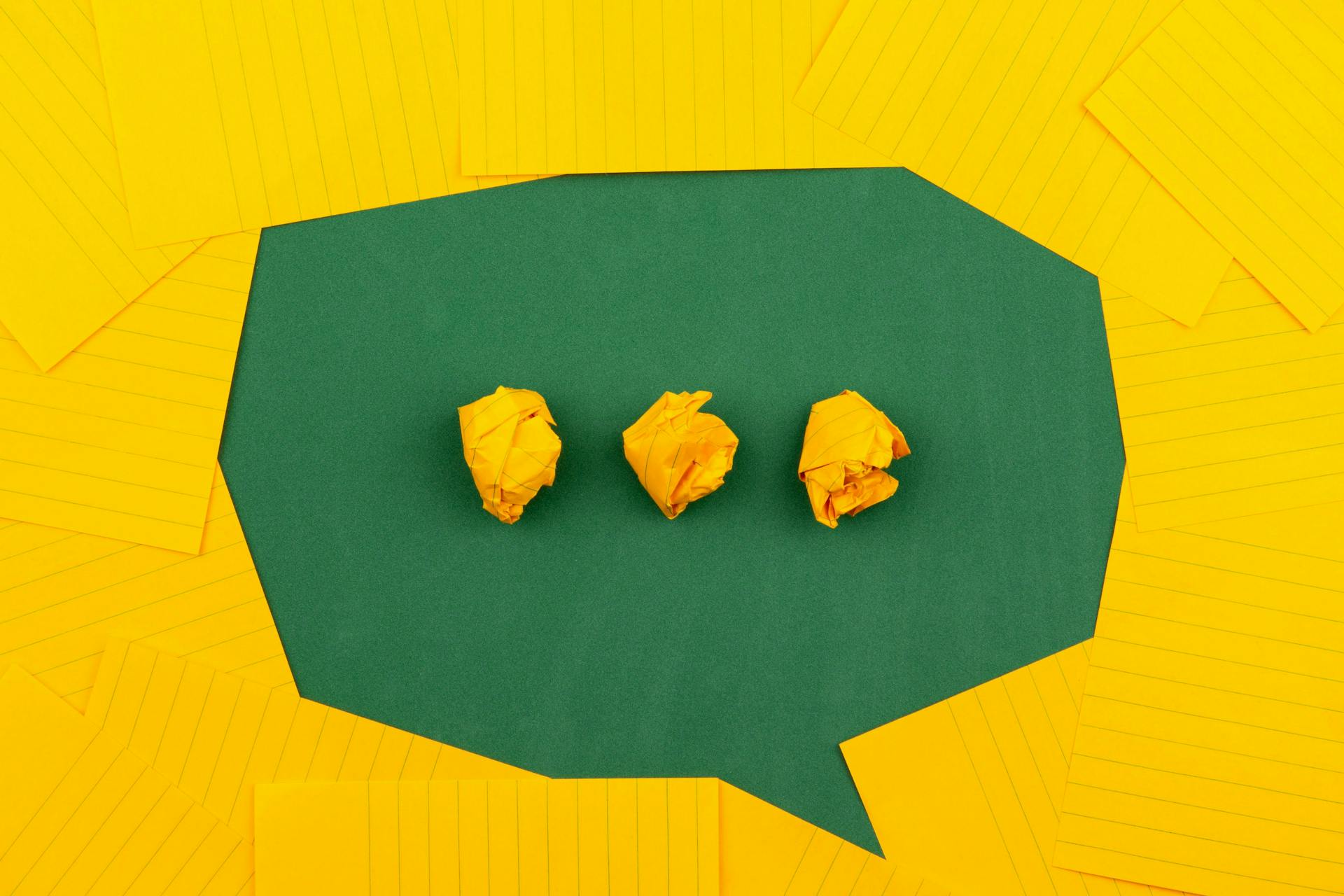 Green speech bubble on yellow background showing three thinking dots made out of craft paper. How to write a communication plan blog post image 