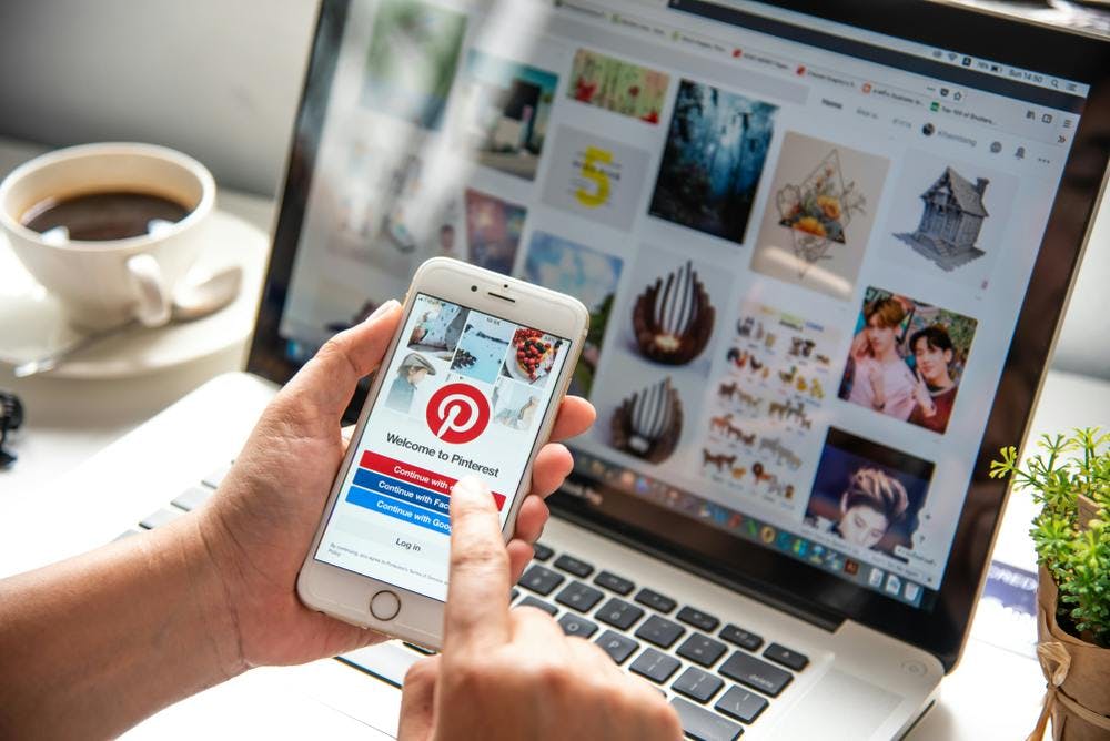 une personne utilise un smartphone affichant la page d'accueil de Pinterest, devant lui se trouve un ordinateur portable et une tasse de café.