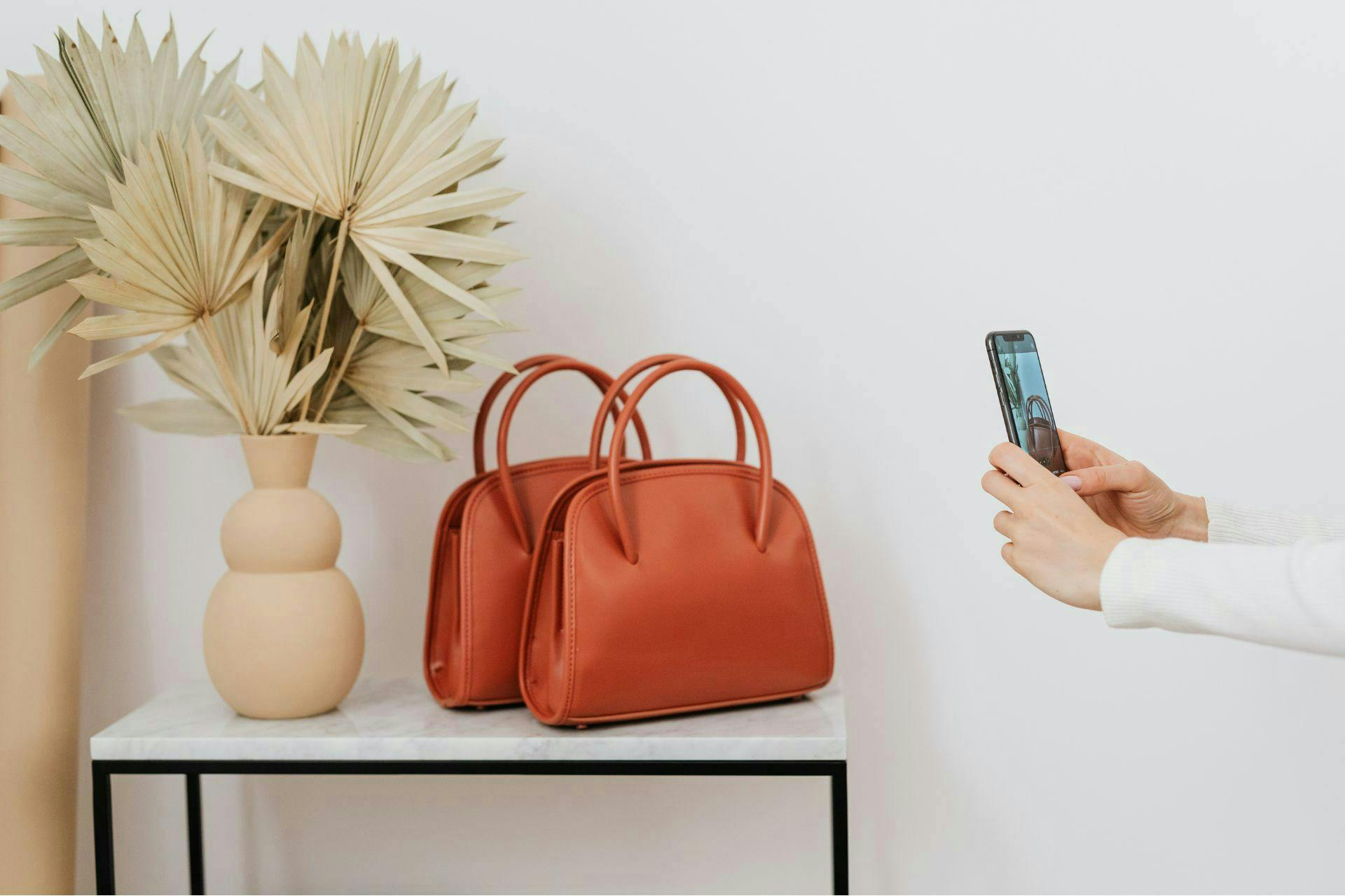 Femme qui prend en photo avec son smartphone des sacs à main sur une table avec un vase derrière