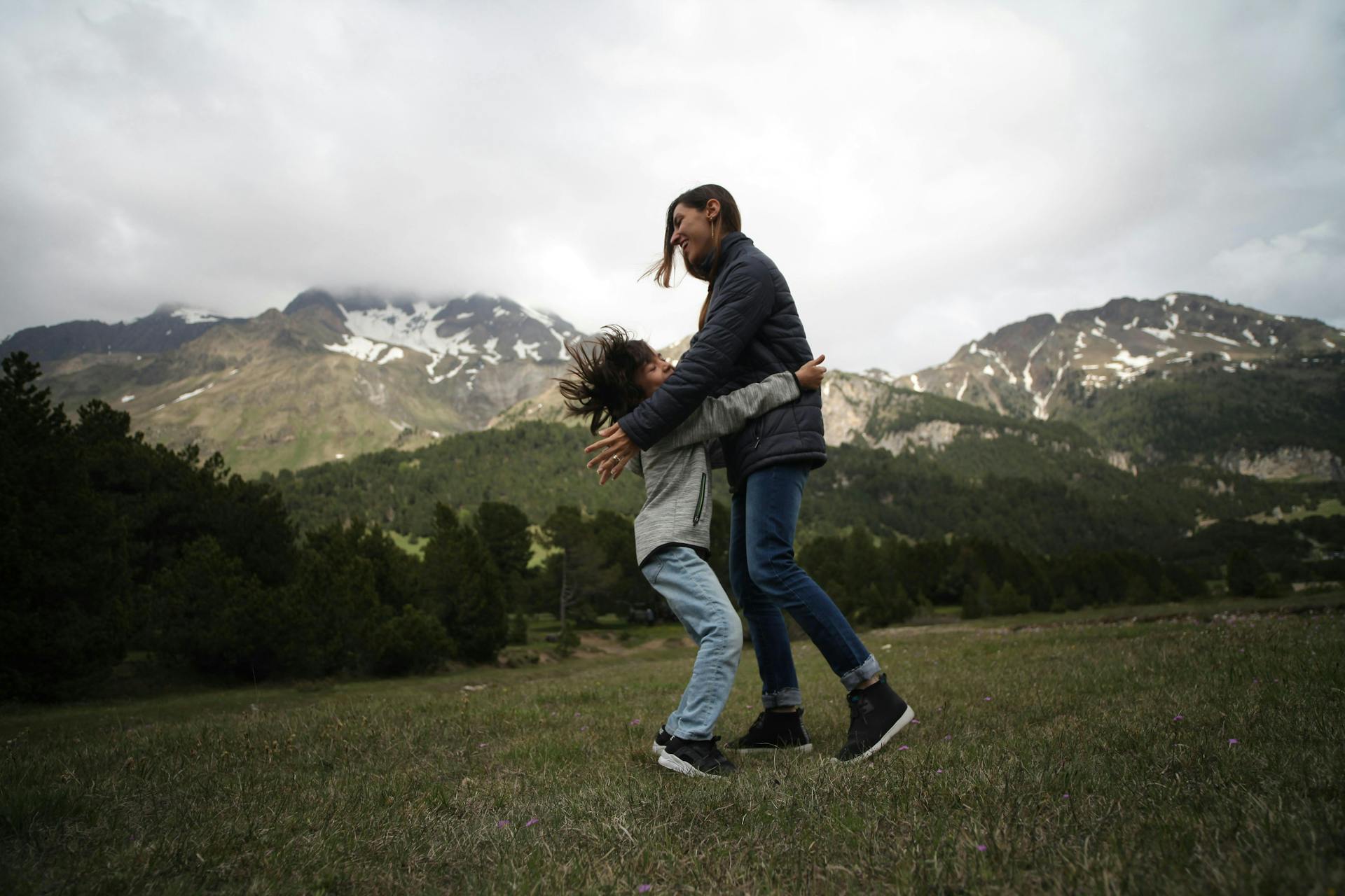Een afbeelding met een kind en een jongen vrouw tegen samen aan het spelen zijn. Op de achtergrond zijn ligt besneeuwde bergen te zien. Een erg authentiek plaatje.