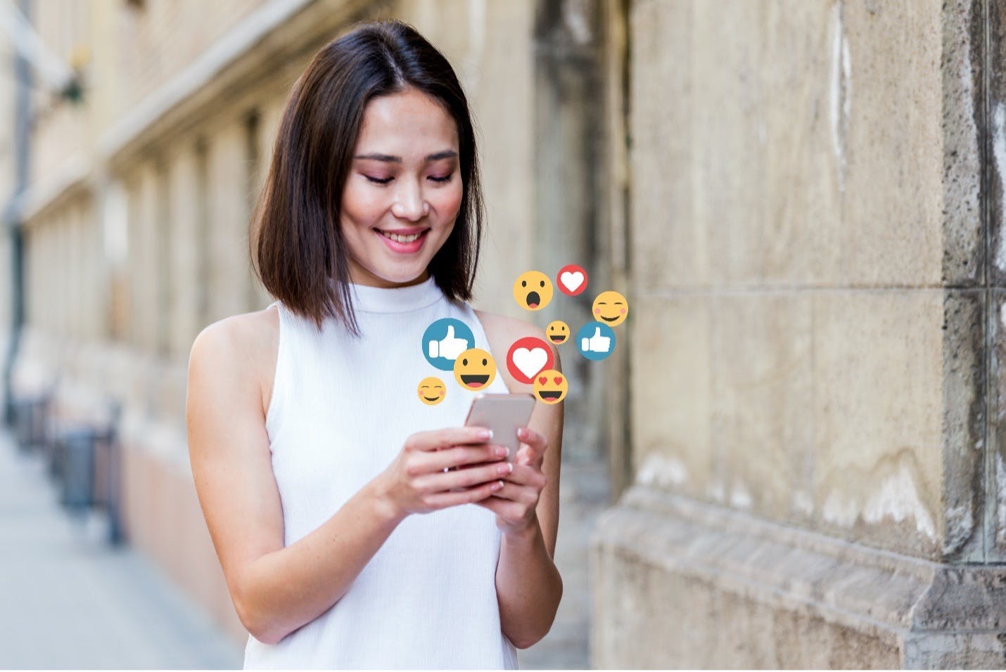 Een vrouw loopt op straat en kijkt op haar mobiel waar verschillende emojis uit komen zweven. Ze zou een social media manager kunnen zijn die engaged met haar community terwijl ze weg is.. 