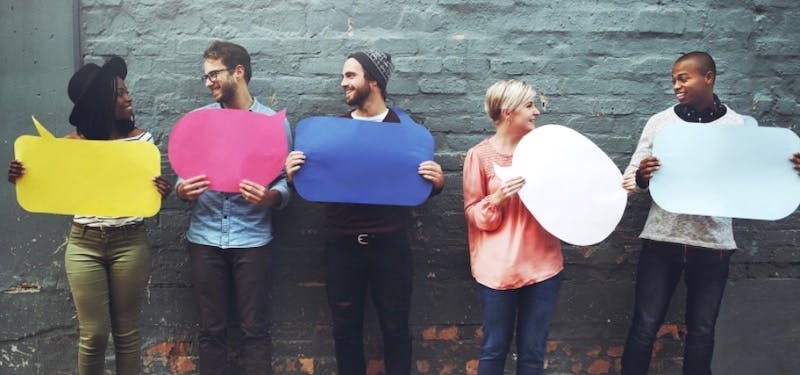 Five young people are standing in front of a brick wall holding colorful speech bubbles made of paper.