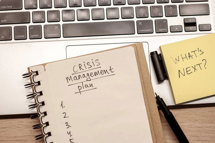 Ein Notizbuch mit der Überschrift "Crises Management Plan" liegt auf einem Laptop.