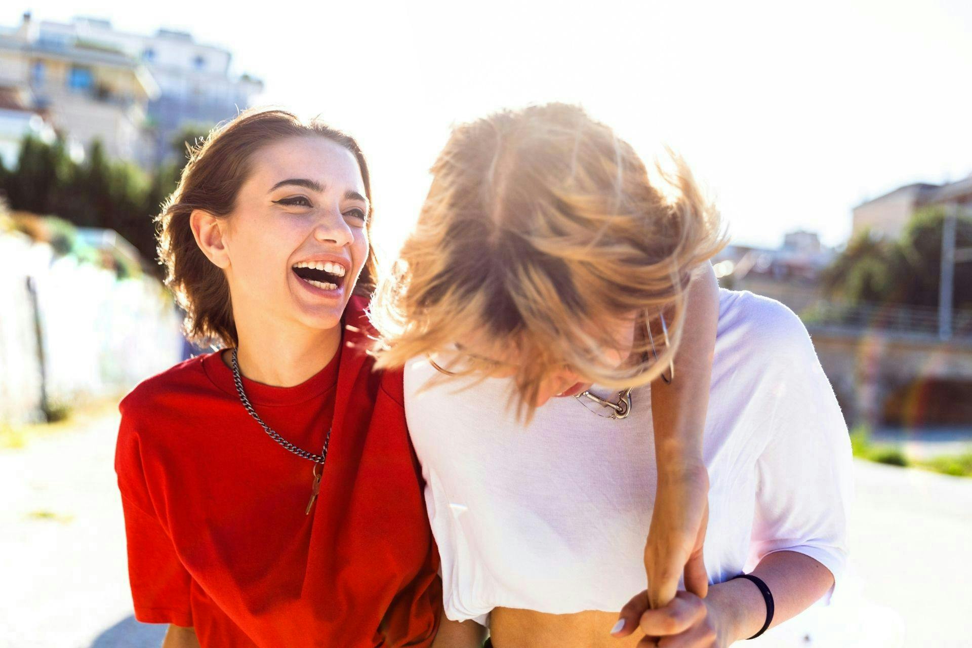 Deux jeunes femmes, une avec un tshirt rouge, l'autre avec un tshirt blanc, dehors, qui rient aux éclats 