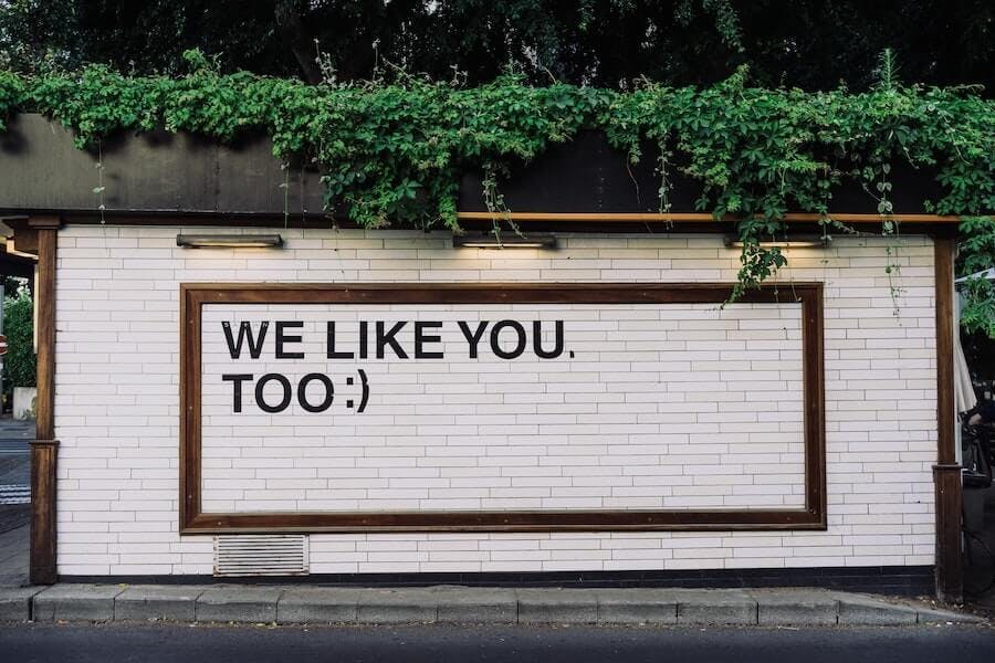 Un mur de briques blanches sur lequel figure le texte "We like you, too :)" (Nous vous aimons aussi). Cet exemple illustre la façon dont les messages marketing peuvent susciter des émotions chez les gens dans le cadre du guérilla marketing.