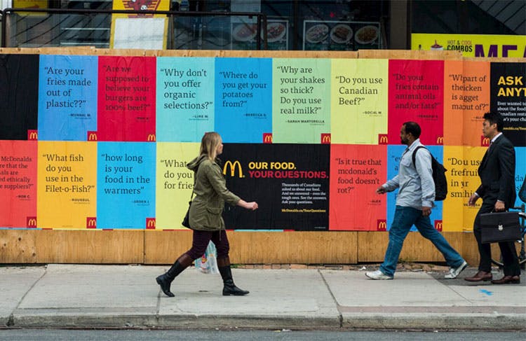 Photo dans une rue - palissade avec des affiches d'une campagne marketing originale de McDonald's. Des personnes marchent dans la rue devant cette campagne. 
